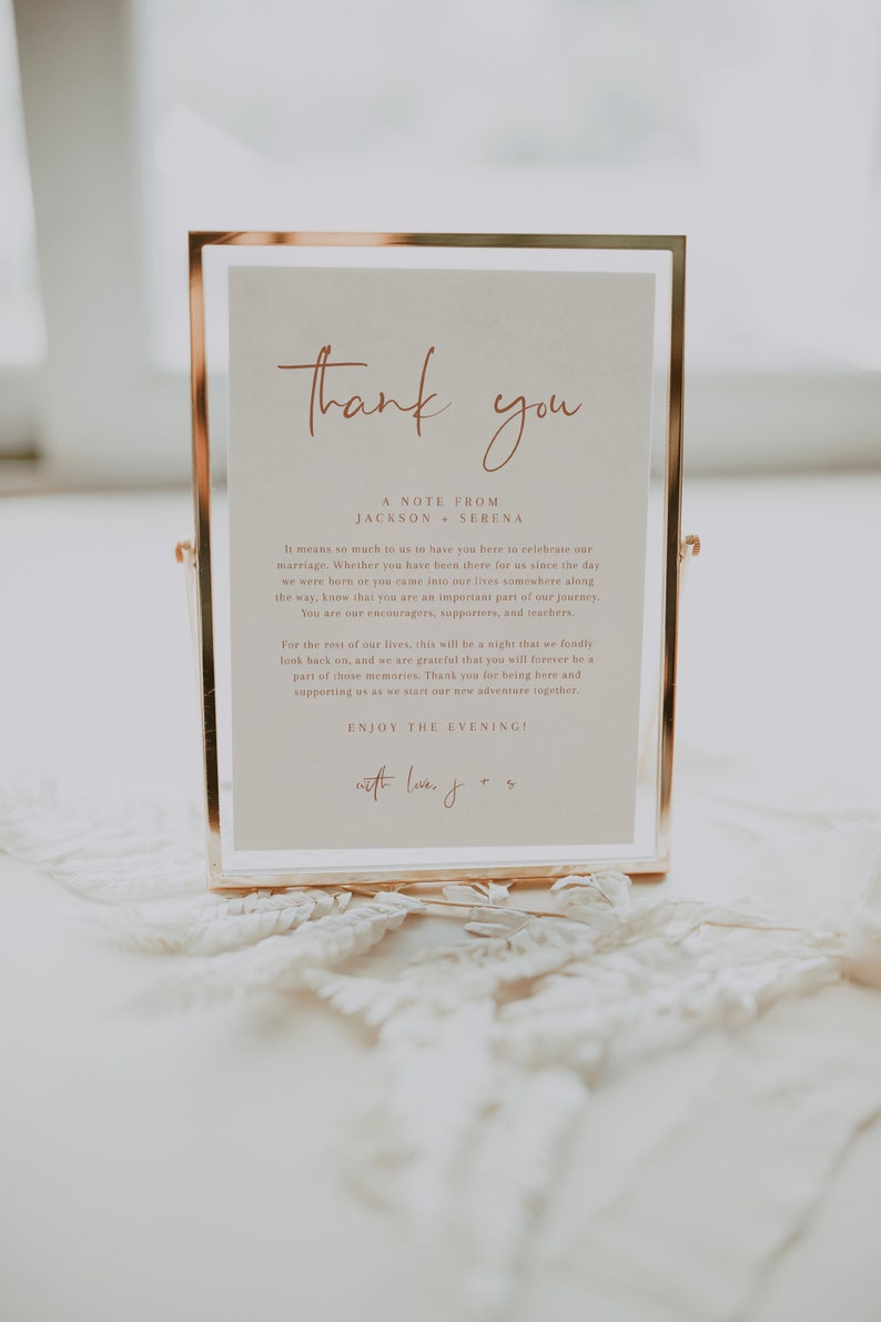 May include: A gold-framed sign with a white background and the text "Thank you" in a cursive font. The sign also includes the text "A Note From Jackson & Serena" and a message expressing gratitude to guests for attending their wedding. The sign is sitting on a white lace surface.