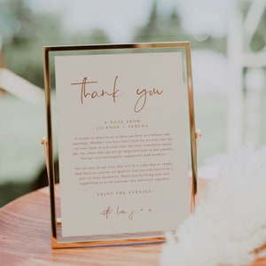 May include: A gold-framed sign with a white background and brown text that reads "Thank you, A Note From Jackson & Serena". The text continues with a message of gratitude to guests for attending their wedding. The sign is on a wooden surface.