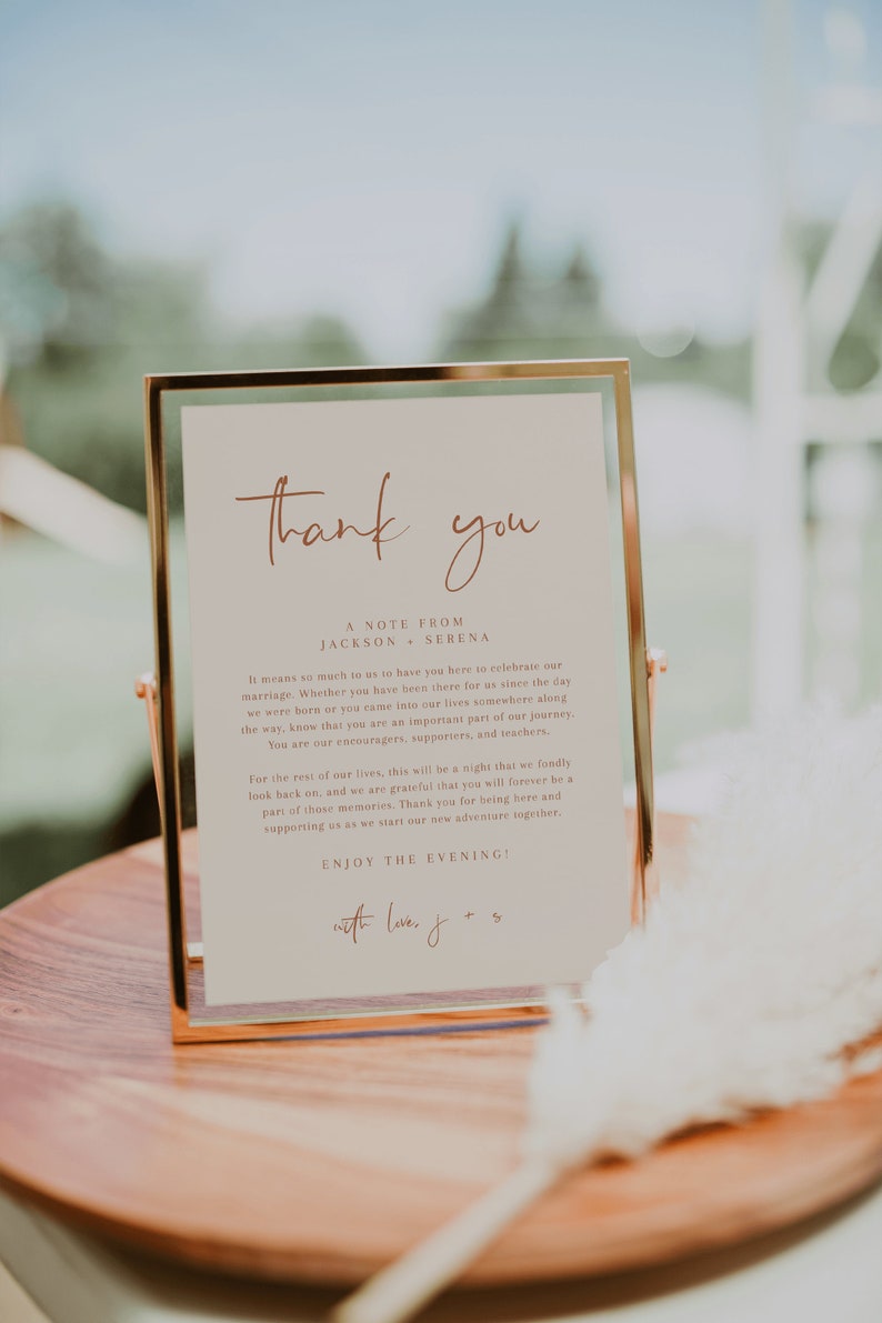 May include: A gold-framed sign with a white background and brown text that reads "Thank you, A Note From Jackson & Serena". The text continues with a message of gratitude to guests for attending their wedding. The sign is on a wooden surface.