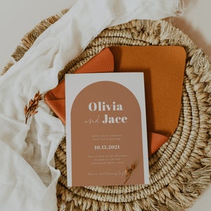 May include: A wedding invitation with a modern design featuring a terracotta-colored arch with the names "Olivia and Jace" in white text. The date "10.15.2021" is printed below the names. The invitation is on a woven placemat with a brown leather envelope.