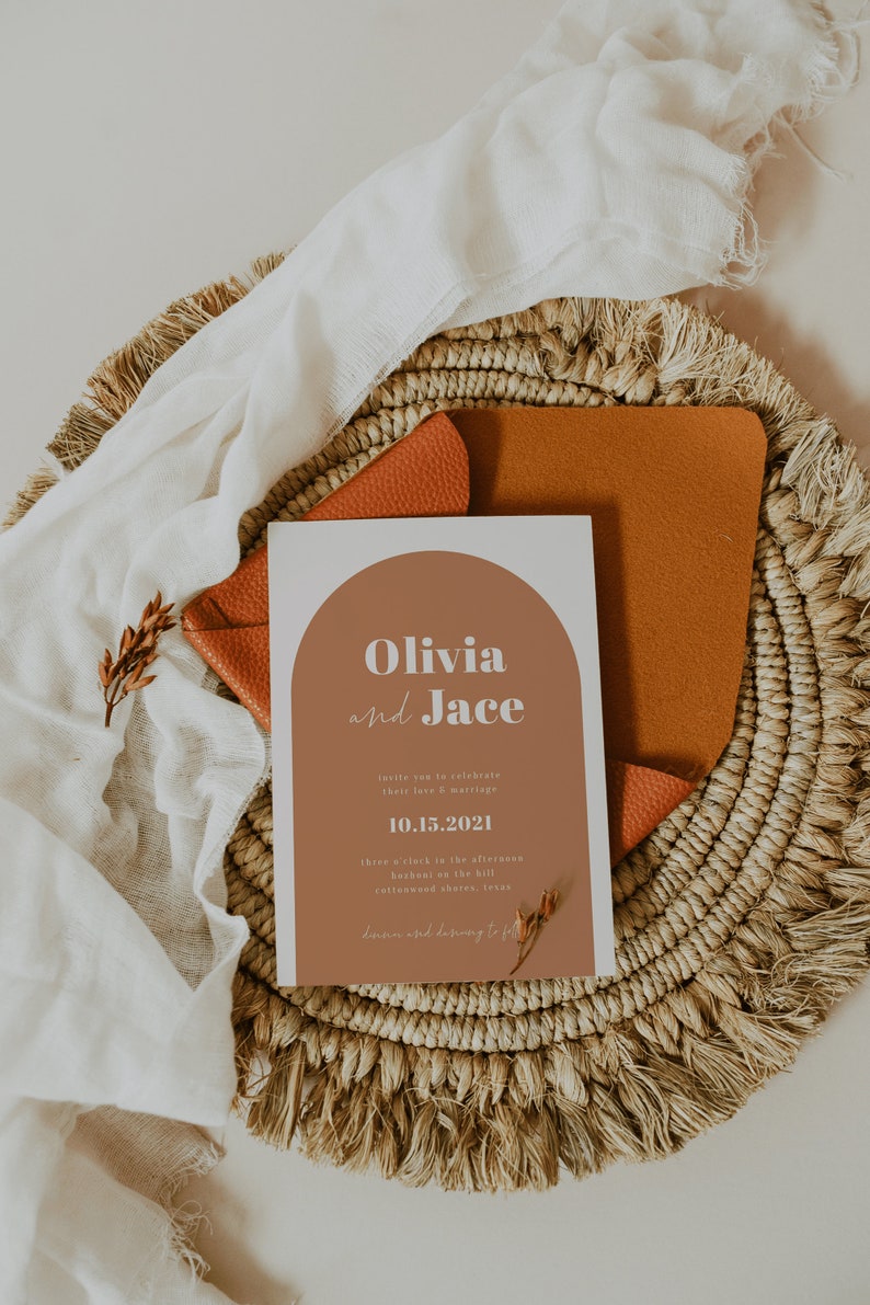May include: A wedding invitation with a modern design featuring a terracotta-colored arch with the names "Olivia and Jace" in white text. The date "10.15.2021" is printed below the names. The invitation is on a woven placemat with a brown leather envelope.