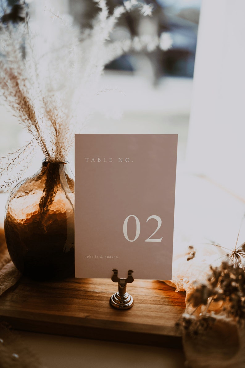May include: A light brown card with the text "TABLE NO. 02" and the text "ophelia & hudson" in white. The card is on a wooden stand with a silver base.