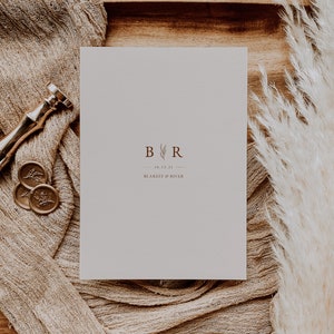 May include: A white card with a brown monogram "B&R" with the date "10.13.23" and the text "BLAKELY & RIVER" printed on it. The card is on a wooden tray with a brown fabric and a white fluffy plant.