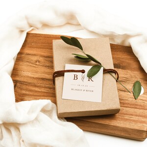 May include: A brown gift box with a white tag tied with brown string. The tag has the initials "B & R" with a small leaf design and the date "10.15.21". The text "BLAKELY & RIVER" is printed below the date.
