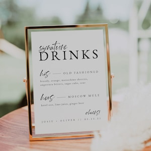 May include: A gold-framed sign with white background and black text. The sign reads "Signature Drinks" and lists two drinks: "his - Old Fashioned" and "hers - Moscow Mule". The sign also includes the text "cheers!" and "JOSIE + OLIVER // 05.15.21".