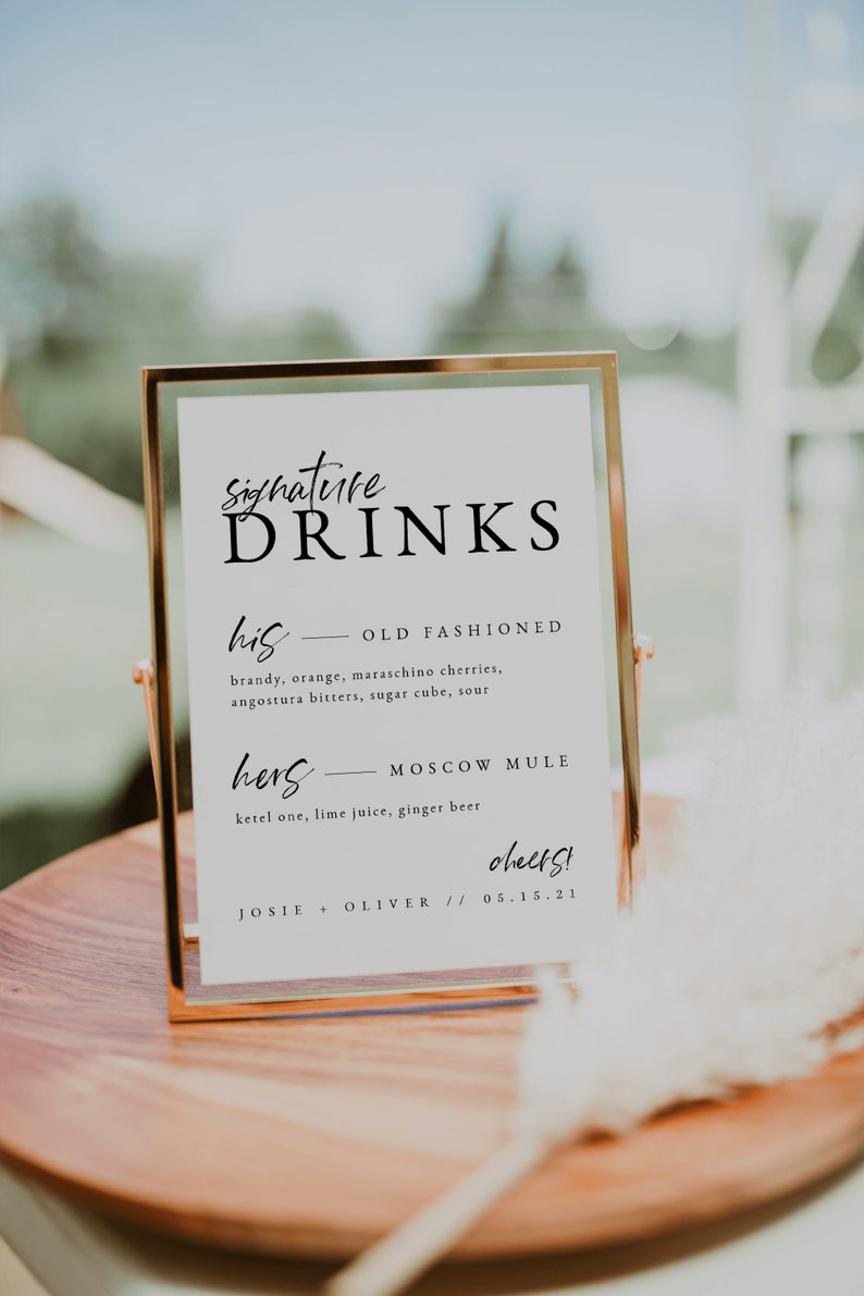 May include: A gold-framed sign with white background and black text. The sign reads "Signature Drinks" and lists two drinks: "his - Old Fashioned" and "hers - Moscow Mule". The sign also includes the text "cheers!" and "JOSIE + OLIVER // 05.15.21".