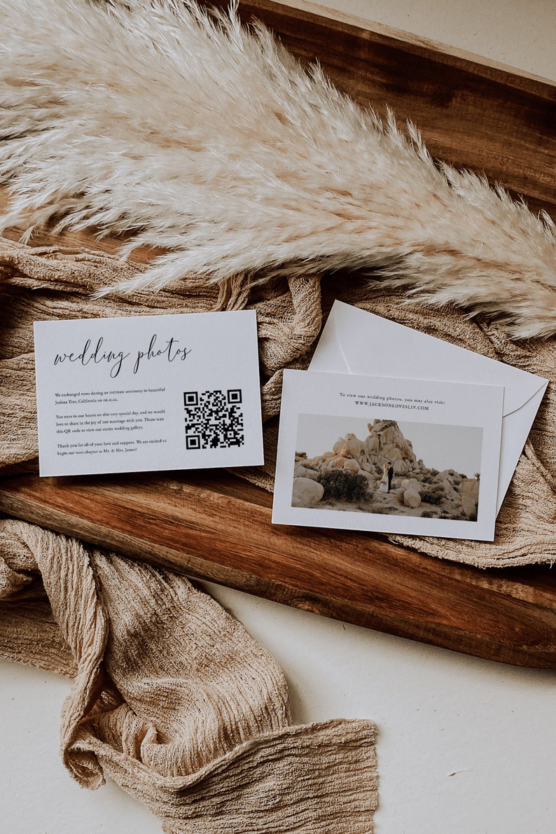 May include: Two white cards on a wooden surface with a tan fabric background. The card on the left has the text "wedding photos" and a QR code. The card on the right has a photo of a couple standing in a desert landscape with the text "To view our wedding photos, you may also visit: www.jacksonloveslife.com".