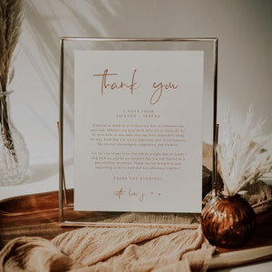 May include: A framed thank you note with a gold frame and a brown background. The note reads "Thank you" in cursive with a note from Jackson and Serena. The note expresses gratitude to guests for attending their wedding and supporting them as they start their new adventure together. The note ends with "Enjoy the evening!" and "with love, J + S".