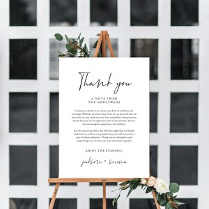 May include: A white sign with black text that reads "Thank you, A Note from the Newlyweds". The text continues with a message of gratitude to guests for attending the wedding. The sign is on a wooden easel with greenery and white flowers.