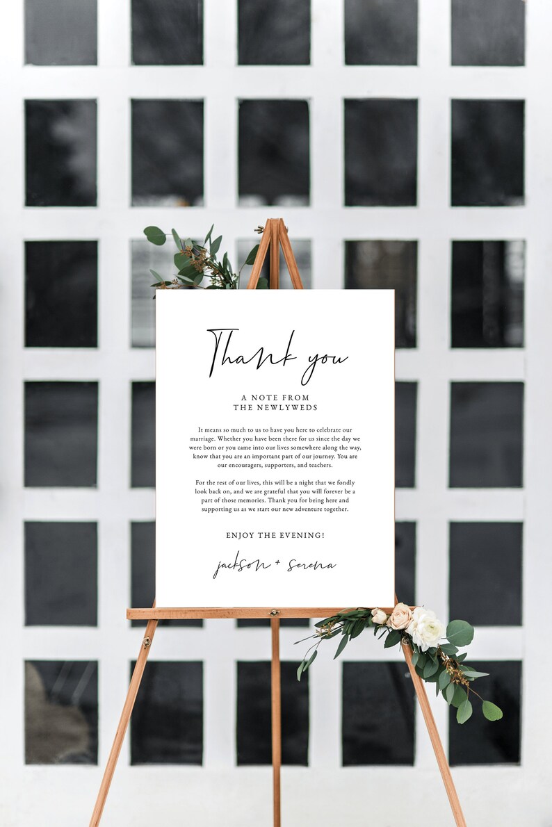 May include: A white sign with black text that reads "Thank you, A Note from the Newlyweds". The text continues with a message of gratitude to guests for attending the wedding. The sign is on a wooden easel with greenery and white flowers.