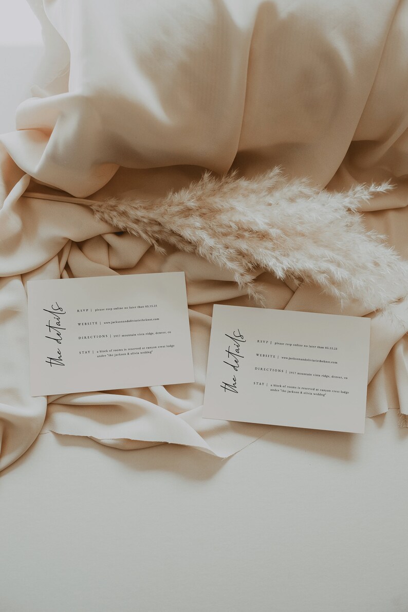 May include: Two white cards with the text "The details" written in black cursive. The cards contain information about an event, including the RSVP, website, directions, and stay. The cards are on a beige fabric background with a dried pampas grass.