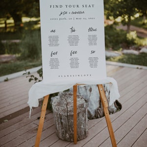 May include: A white sign with black text on an easel. The sign reads "Find Your Seat Josie + Cameron Estes Park, CO | May 15, 2021" and lists names under the headings "One", "Two", "Three", "Four", "Five", and "Six".