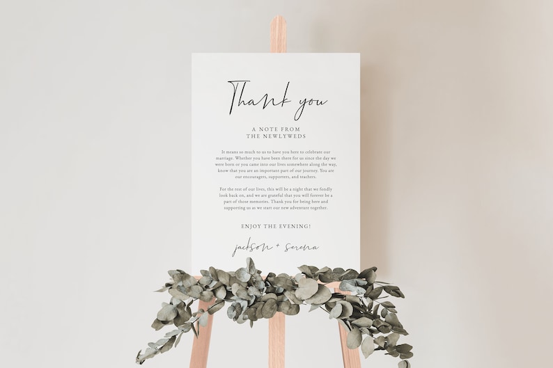 May include: A white sign with black text that reads "Thank you, A note from the newlyweds." The text continues with a message of gratitude to guests for attending the wedding. The sign is on a wooden easel with greenery around it.