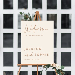 May include: Welcome sign with a cream background and brown text. The sign reads "Welcome to the wedding of Jackson and Sophie, October 15, 2021". The sign is on an easel with greenery and flowers.