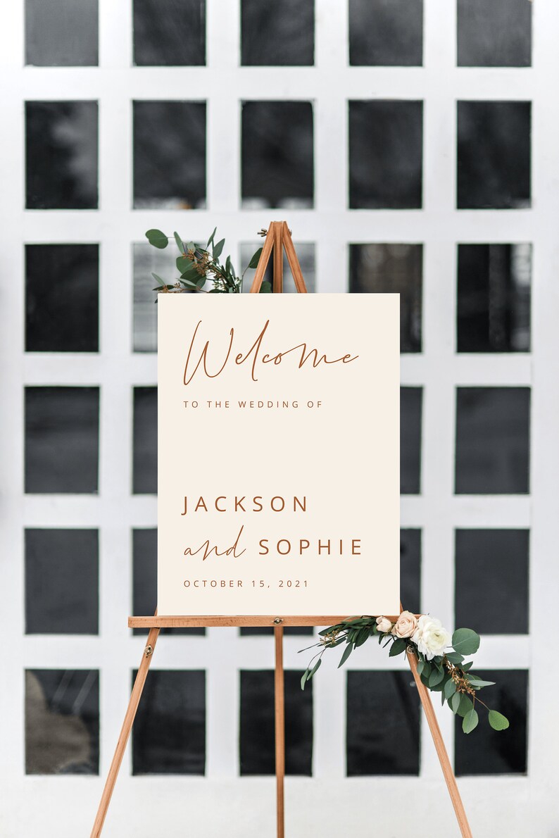 May include: Welcome sign with a cream background and brown text. The sign reads "Welcome to the wedding of Jackson and Sophie, October 15, 2021". The sign is on an easel with greenery and flowers.