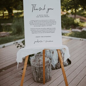 May include: A white sign with black text on an easel. The sign says "Thank you, A Note From The Newlyweds". The text continues with a message of gratitude to guests for attending the wedding. The sign ends with "Enjoy the Evening! Jackson + Serena".
