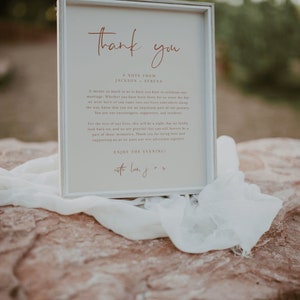 May include: A white framed sign with a thank you message for guests attending a wedding. The sign reads "Thank you" in a cursive font with the text "A note from Jackson + Serena" below. The message expresses gratitude to guests for their presence and support. The sign also includes the text "Enjoy the evening! with love, J + S".