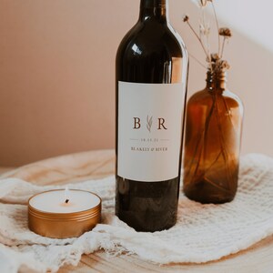 May include: A wine bottle with a white label that says "B R" with a leafy design and the date "10.15.21" and the text "BLAKELY & RIVER". The bottle is on a wooden surface with a white cloth and a small gold candle.