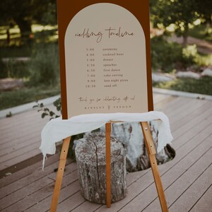 May include: A brown and beige wedding timeline sign with a rounded arch at the top. The sign lists the wedding events with times, including the ceremony, cocktail hour, dinner, speeches, first dance, cake cutting, late night pizza, and send off. The sign also includes the names Kinsley and Teegan.