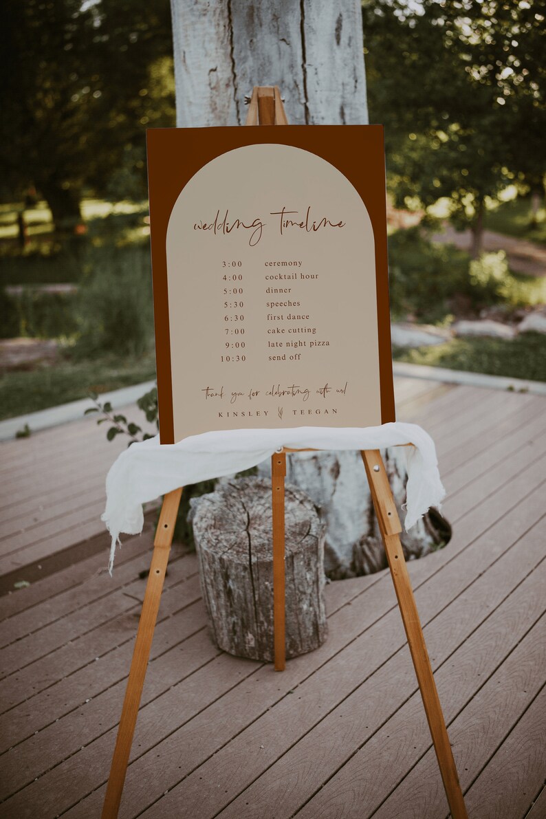 May include: A brown and beige wedding timeline sign with a rounded arch at the top. The sign lists the wedding events with times, including the ceremony, cocktail hour, dinner, speeches, first dance, cake cutting, late night pizza, and send off. The sign also includes the names Kinsley and Teegan.