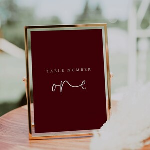 Puede incluir: Un letrero de n&uacute;mero de mesa con marco dorado, fondo rojo oscuro y texto blanco que dice "TABLE NUMBER one".