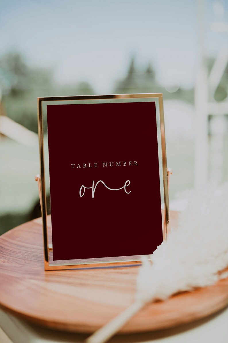 Puede incluir: Un letrero de n&uacute;mero de mesa con marco dorado, fondo rojo oscuro y texto blanco que dice "TABLE NUMBER one".