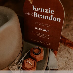 May include: A brown and white wedding invitation with an arched top. The invitation reads "Kenzie and Brandon" and includes the date "03.27.2021". The invitation is sitting on a brown hat with two ring boxes.