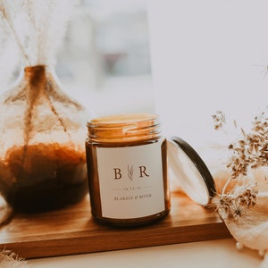 May include: A brown glass candle jar with a white label. The label has a monogram "B.R." with a small leaf design and the text "10.15.21 - BLAKELY & RIVER".