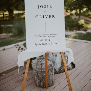 May include: A white sign with black text announcing the wedding of Josie and Oliver. The sign says "Josie + Oliver are delighted to welcome you to their wedding 08-07-21 Laguna Beach, California."
