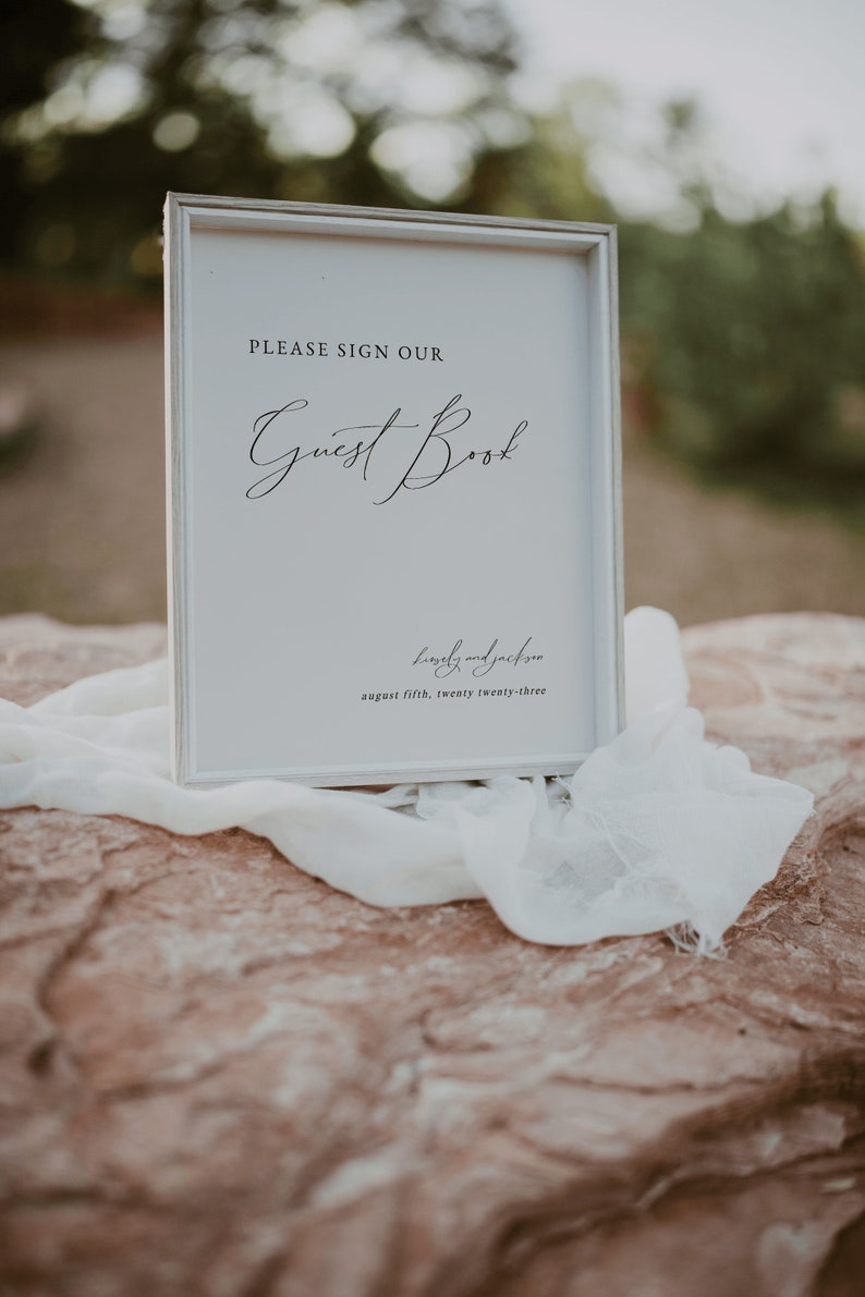 May include: White sign with a black frame, reading "PLEASE SIGN OUR Guest Book Lovely and Jackson August fifth, twenty twenty-three"