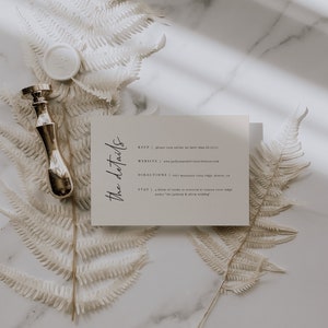May include: A white card with the details for a wedding. The card has the text "The details" in a script font. The text on the card includes the RSVP, website, directions, and stay information. The card is on a white marble surface with dried ferns.