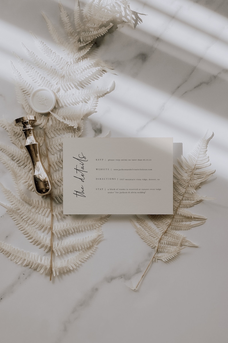 May include: A white card with the details for a wedding. The card has the text "The details" in a script font. The text on the card includes the RSVP, website, directions, and stay information. The card is on a white marble surface with dried ferns.