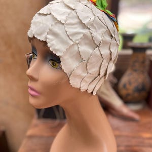 Puede incluir: Un sombrero blanco con bordes festoneados y detalles florales coloridos. El sombrero está hecho de formas superpuestas en forma de pétalos y se exhibe en una cabeza de maniquí. Las flores son amarillas, rosas y verdes.