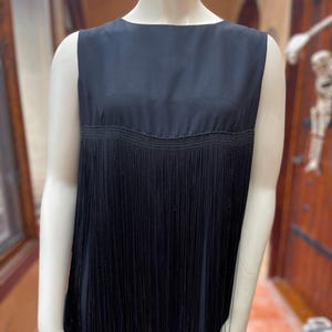 Puede incluir: Vestido negro sin mangas con cuello redondo y detalles de flecos. El vestido presenta un corpiño liso y una falda con flecos. Los flecos son largos y cuelgan de la cintura. El vestido se exhibe en un maniquí.