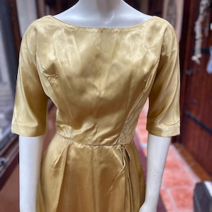 Peut inclure: Robe dorée vintage avec encolure bateau et manches trois-quarts. La robe présente un corsage ajusté et une jupe plissée, créant une silhouette classique. Le tissu a un éclat subtil, suggérant du satin ou de la soie. La robe est présentée sur un mannequin.