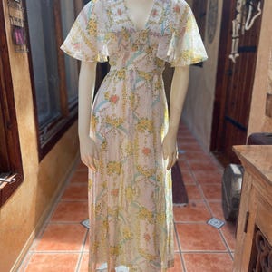 May include: A floor-length dress featuring a V-neckline, flutter sleeves, and a smocked bodice. The fabric is white with a repeating pattern of yellow flowers, orange floral accents, and light blue ribbon-like stripes throughout the garment.