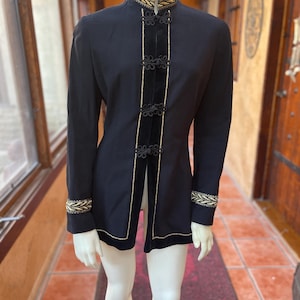 May include: Black jacket with gold trim and accents. The jacket features a high collar and a velvet panel down the front, secured with decorative buttons. Gold trim adorns the collar, cuffs, and edges, creating a sophisticated look. The jacket is displayed on a mannequin.