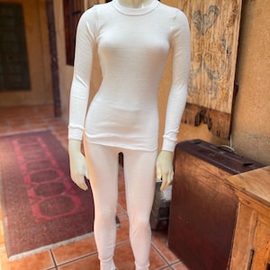 May include: A white thermal underwear set displayed on a mannequin. The set includes a long-sleeved crew neck top and matching leggings. The fabric appears to be a soft, knit material. The mannequin is on a metal stand, set against a backdrop of a rustic interior.