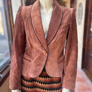 May include: A brown suede blazer with a notched lapel and a single button closure. The blazer has a tailored fit and features a pocket on each side. The mannequin is also wearing a skirt with a geometric pattern.