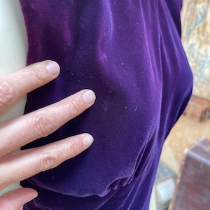 May include: A sleeveless, deep purple velvet dress. The dress has a scoop neckline trimmed with a silver-coloured embellishment. The fabric is gathered at the waist, creating a flattering silhouette. The dress is made of a soft, luxurious velvet material.