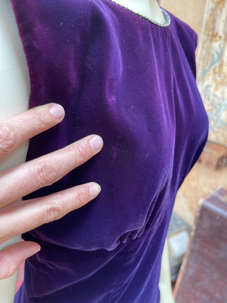 May include: A sleeveless, deep purple velvet dress. The dress has a scoop neckline trimmed with a silver-coloured embellishment. The fabric is gathered at the waist, creating a flattering silhouette. The dress is made of a soft, luxurious velvet material.