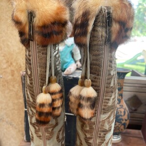 May include: A pair of tall, decorative boots with a tan and brown color scheme. The boots feature a textured, faux-snakeskin pattern and intricate embroidered designs. Each boot has a fluffy, fur-trimmed top and decorative tassels.