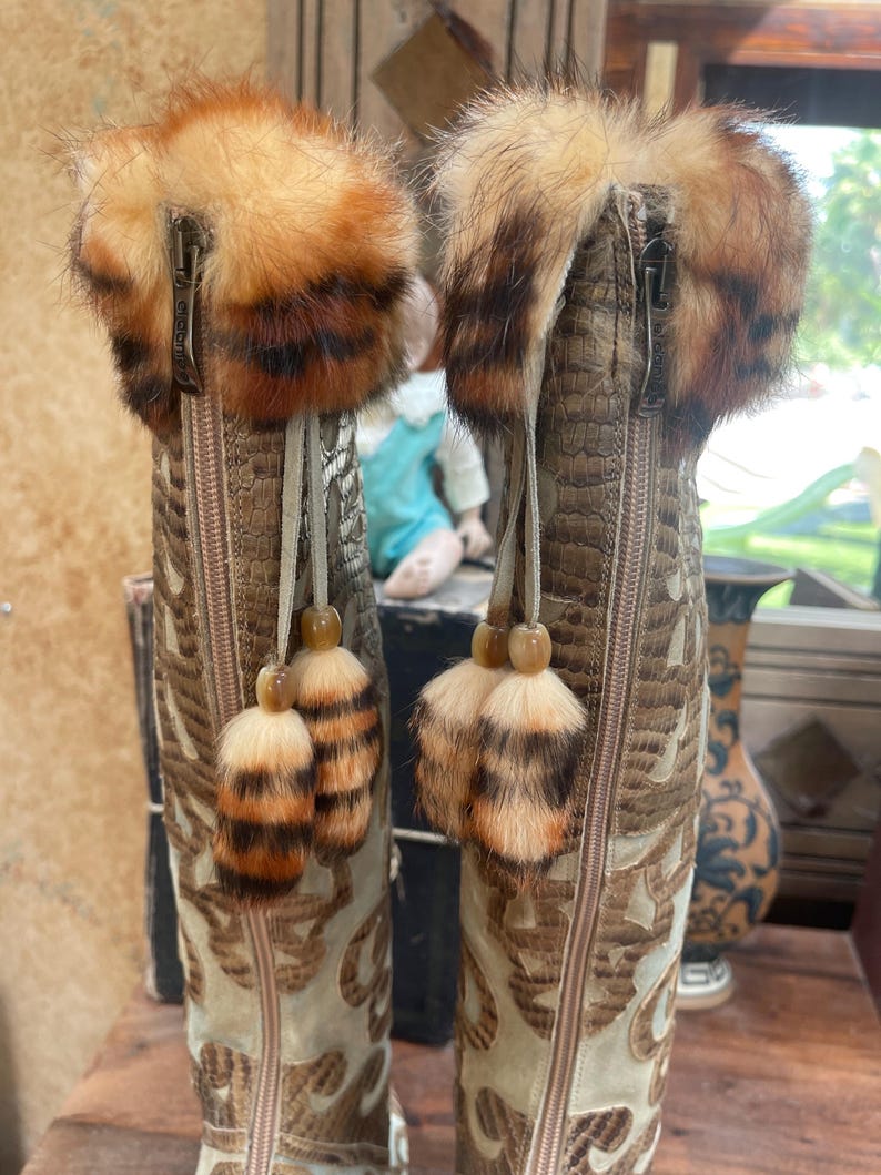 May include: A pair of tall, decorative boots with a tan and brown color scheme. The boots feature a textured, faux-snakeskin pattern and intricate embroidered designs. Each boot has a fluffy, fur-trimmed top and decorative tassels.