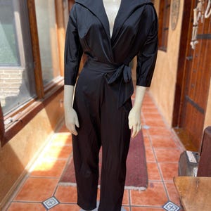 May include: A black jumpsuit with a wrap-style bodice and a tie belt. The jumpsuit has three-quarter sleeves and wide-leg pants. The outfit is displayed on a mannequin.