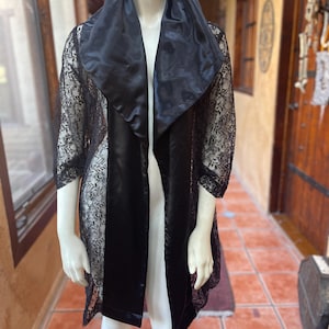 May include: A black lace and satin robe displayed on a mannequin. The robe features a wide satin collar and sleeves with intricate lace detailing. The robe is open in the front, revealing the mannequin's form. The background includes a doorway and windows.