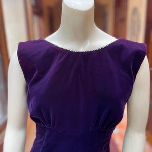 May include: A sleeveless, deep purple velvet dress on a mannequin. The dress has a scoop neckline with a delicate trim and a fitted waist. The fabric has a soft, luxurious appearance, and the dress is likely vintage or retro.