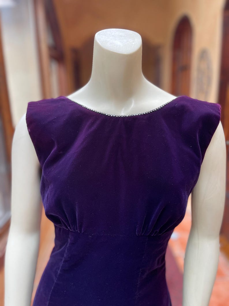 May include: A sleeveless, deep purple velvet dress on a mannequin. The dress has a scoop neckline with a delicate trim and a fitted waist. The fabric has a soft, luxurious appearance, and the dress is likely vintage or retro.
