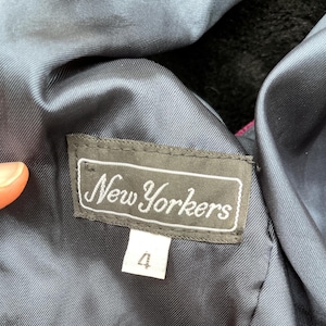 May include: A close-up of a clothing label reading "New Yorkers" in a cursive font. The label is dark grey with a white border and text. Below is a white tag with the number "4". The garment's lining is a dark blue.