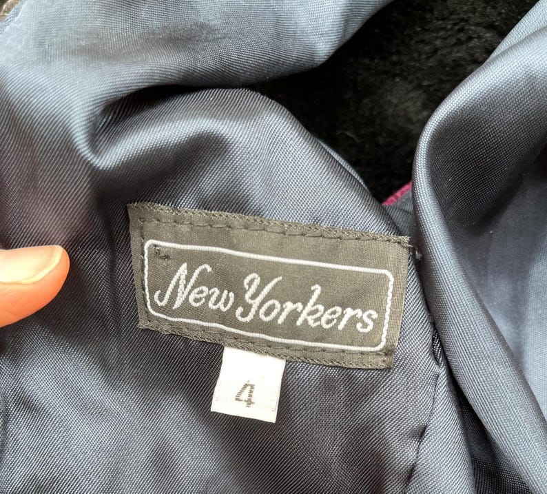 May include: A close-up of a clothing label reading "New Yorkers" in a cursive font. The label is dark grey with a white border and text. Below is a white tag with the number "4". The garment's lining is a dark blue.