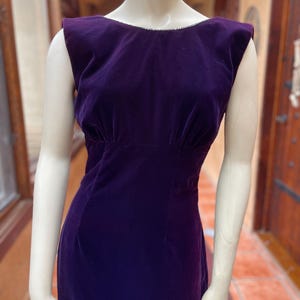 May include: A sleeveless, floor-length, deep purple velvet dress. The dress has a scoop neckline with a delicate trim and a fitted bodice that flares out at the waist. The dress is displayed on a mannequin.